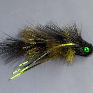 May include: A handmade fishing fly with a black head and body, accented with olive and black feathers. The fly features a bright green eye and neon yellow accents, designed to attract fish.