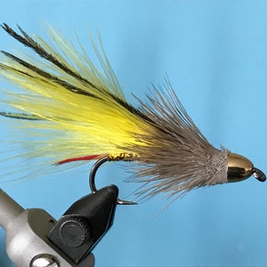 Puede incluir: Un señuelo de pesca con mosca amarillo y gris con una cabeza de cuentas doradas y una cola roja.