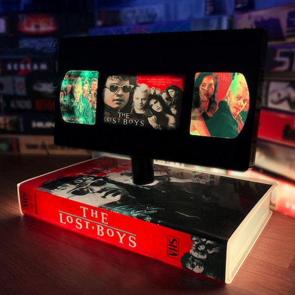May include: A black VHS tape lamp featuring The Lost Boys movie imagery. The lamp shade displays three illuminated panels with scenes from the film, while the base is a red VHS tape case with the text The Lost Boys printed on the side.