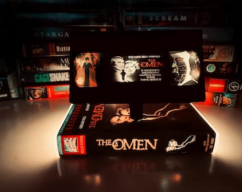 May include: A black VHS tape of "The Omen" movie, with a glowing light illuminating the title and cover art. The tape is displayed in front of a collection of other VHS tapes, creating a nostalgic horror movie theme.