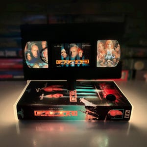 May include: A unique, illuminated VHS tape lamp featuring the movie "Fifth Element." The lamp showcases the VHS tape's cover art and the movie's title, with the tape itself serving as the base and the illuminated screen.