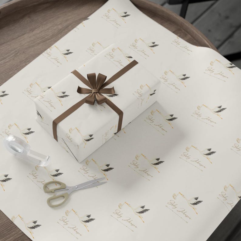 May include: White gift wrap with a repeating pattern of a gold frame with the text "Baby Shower" and a black and white illustration of a stork with feathers. A brown ribbon is tied around a white gift box.