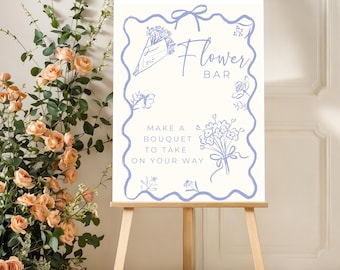 May include: A wooden easel displays a sign with the words "Flower Bar" in blue script. The sign has a light background with blue floral illustrations and the text "Make a bouquet to take on your way."