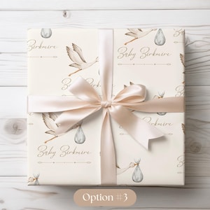 May include: A white gift wrap with a beige ribbon. The wrap features a repeating pattern of storks carrying baby bundles and the text "Baby Birkmire".