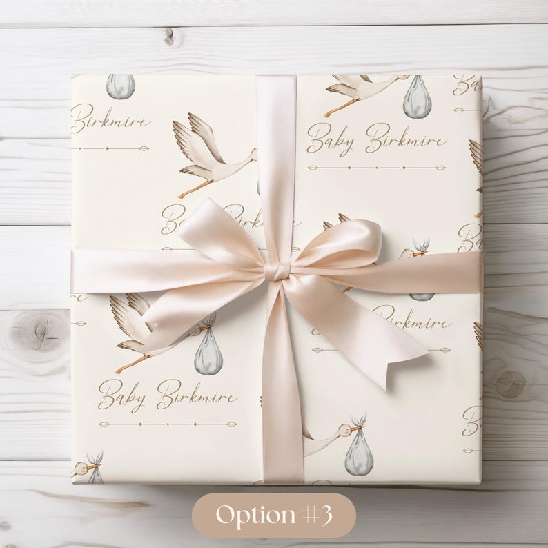 May include: A white gift wrap with a beige ribbon. The wrap features a repeating pattern of storks carrying baby bundles and the text "Baby Birkmire".