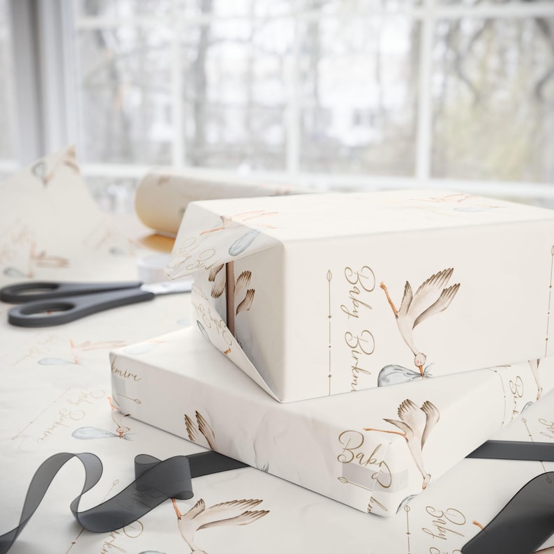 May include: White gift wrap with a stork carrying a baby in a bag. The wrap has a gold inscription that says "Baby Birthday".