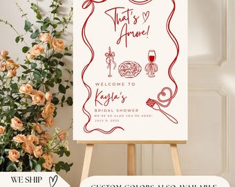 以下が含まれることがあります： イーゼルに立てられたクリーム色の看板。赤い文字とイラストで「That's Amore!」と「Kayla's Bridal Showerへようこそ」と書かれています。食べ物のイラストと日付が含まれています。看板は赤い縁取りがされています。