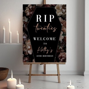 May include: A decorative sign on an easel with the text "RIP twenties WELCOME TO Kathy's 30TH BIRTHDAY" in white and rose gold lettering. The background is a floral pattern. Lit candles and a woven bowl are in the foreground.