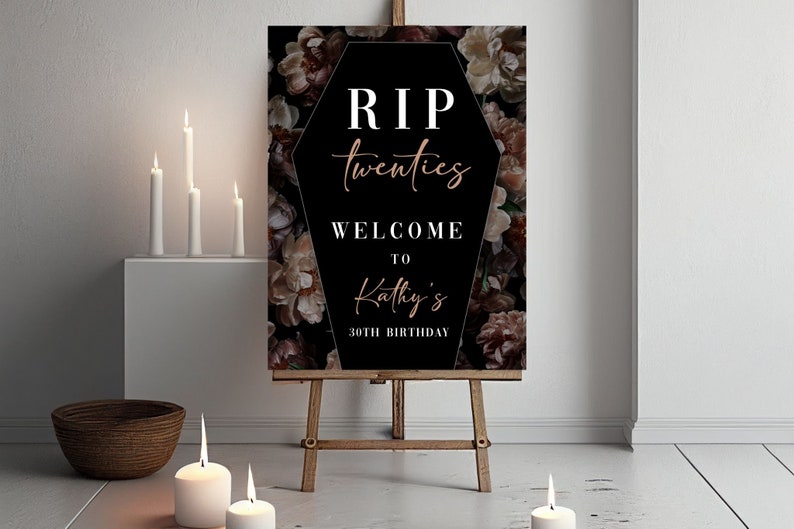May include: A decorative sign on an easel with the text "RIP twenties WELCOME TO Kathy's 30TH BIRTHDAY" in white and rose gold lettering. The background is a floral pattern. Lit candles and a woven bowl are in the foreground.