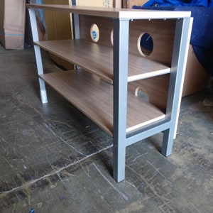 May include: A light brown wooden TV stand with two shelves and silver metal legs. The stand has several round cutouts on the back panel.