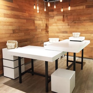 May include: A white L-shaped table with black metal legs. The table is made of wood and has a white top. The table is in a room with wood paneling on the walls. There are light bulbs hanging from the ceiling.