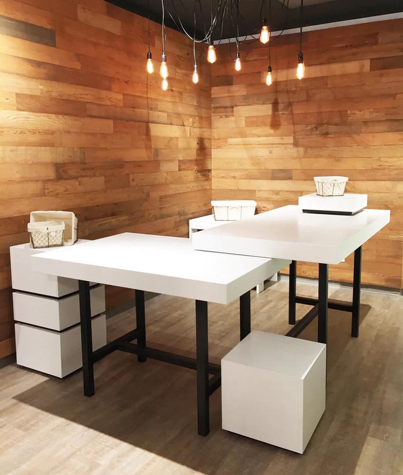 May include: A white L-shaped table with black metal legs. The table is made of wood and has a white top. The table is in a room with wood paneling on the walls. There are light bulbs hanging from the ceiling.