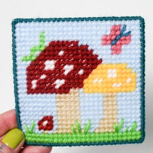 Puede incluir: Una pieza cuadrada bordada a punto de cruz con dos setas, una roja con lunares blancos y otra amarilla, sobre un fondo azul claro. Una mariposa rosa y una flor roja también están bordadas en el diseño, con hierba verde en la parte inferior.