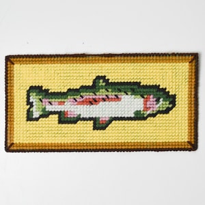 Puede incluir: Un tapiz rectangular hecho a mano con una trucha arcoíris. El pez está detallado con verde, rosa y blanco, sobre un fondo amarillo. La pieza está enmarcada con un borde marrón.