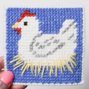 Puede incluir: Un diseño de punto de aguja cuadrado con una gallina blanca con cresta roja y pico amarillo, sobre un fondo azul. La gallina está rodeada de puntadas amarillas y grises, creando un diseño encantador y rústico.