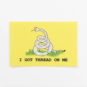 Puede incluir: Un fondo amarillo con una serpiente blanca enroscada en un círculo con la lengua fuera. La serpiente está sobre un lecho de hierba verde. El texto "I GOT THREAD ON ME" está debajo de la serpiente.