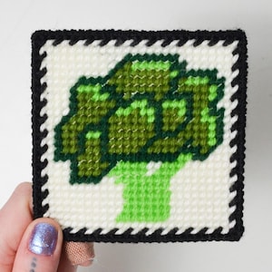 Puede incluir: Posavasos cuadrado hecho a mano con punto de cruz, con un diseño de brócoli verde sobre un fondo blanco. El posavasos tiene un borde negro. El diseño está hecho con hilos verdes.