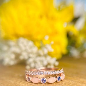 Könnte beinhalten: Zwei zarte Ringe mit einer Roségold-Oberfläche. Der obere Ring hat eine Reihe kleiner, klarer Edelsteine. Der untere Ring hat eine Reihe ovaler, rosafarbener Edelsteine mit blauen Edelsteinen dazwischen.