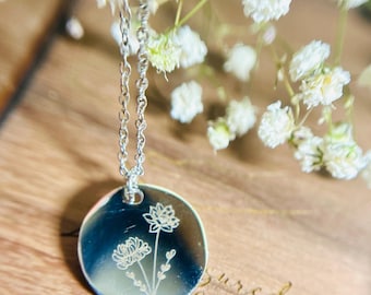 May include: A silver necklace with a round pendant featuring an engraved floral design. The pendant has two flowers with stems and leaves.