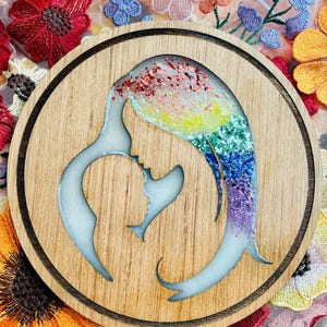 Könnte beinhalten: Ein runder Untersetzer aus Holz mit einer weißen Silhouette einer Mutter und eines Kindes. Die Silhouette ist mit Regenbogen-Glitzerharz gefüllt.