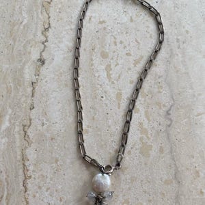 May include: A silver-toned chain necklace with a pendant featuring a large, white pearl and smaller, light-colored beads. The necklace is displayed on a light-colored, textured surface.