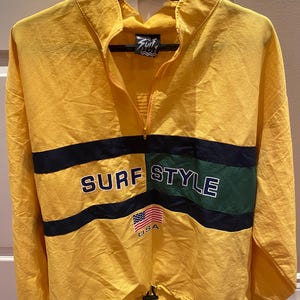 May include: Yellow windbreaker with a half-zip, black and green stripes, and navy blue "SURF STYLE" text. Includes a small American flag and "USA". The jacket hangs on a black hanger. Measurements are not available.