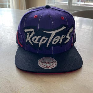 May include: A purple and black Raptors baseball cap with an adjustable fit. The word "Raptors" is in white script across the front. The brim is black, and the cap has red accents. A circular tag is visible.