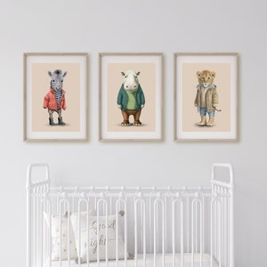 Puede incluir: Tres ilustraciones enmarcadas de animales con ropa. Los animales son una cebra, un hipopótamo y un león. La cebra lleva una chaqueta roja, el hipopótamo lleva una chaqueta verde y el león lleva una chaqueta azul. Los marcos son todos del mismo tamaño y están colgados en una pared blanca. Hay una cuna blanca con un colchón blanco y un caballo de balancín blanco en primer plano.