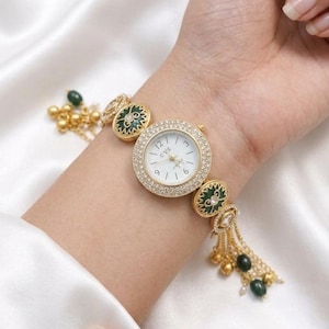 May include: Gold-coloured wristwatch with a white face and crystal-studded bezel. The watch features green and gold decorative elements and a chain with dangling beads. The watch is on a wrist against a white satin background.