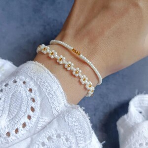 May include: Two beaded bracelets. One bracelet features small white beads with gold accents in a daisy pattern. The other bracelet is a simple strand of white beads with a gold accent. The bracelets are worn on a wrist.
