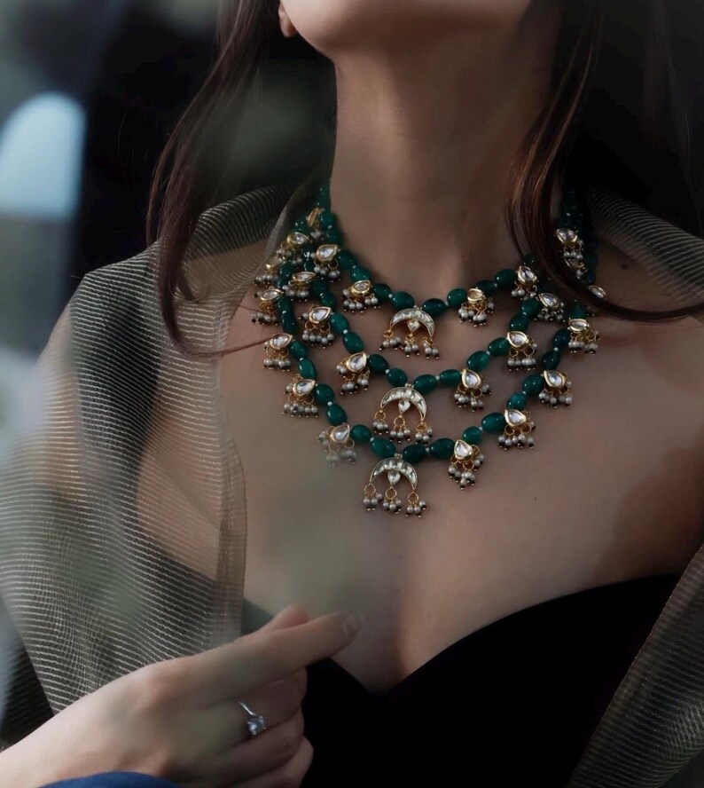 Op de afbeelding: Een gelaagde smaragdgroene en gouden ketting met parel accenten en halve maan bedels.
