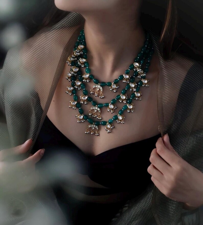 Op de afbeelding: Een gelaagde smaragdgroene en gouden ketting met traanvormige kralen en decoratieve accenten.