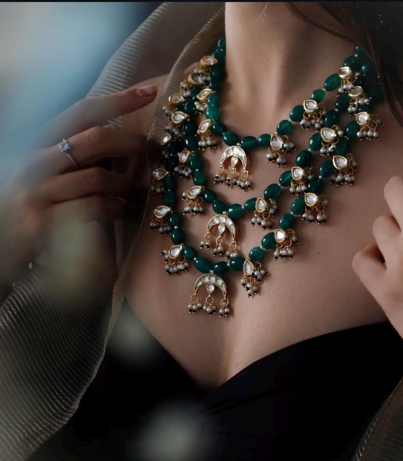 Op de afbeelding: Een gelaagde groene en gouden ketting met traanvormige kralen en halvemaan hangers. De ketting is gemaakt van kralen, metaal en stenen.