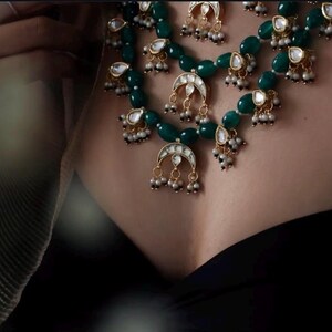 Op de afbeelding: Een gelaagde ketting met groene kralen, gouden accenten en parel-achtige druppels. De ketting heeft een halve maan hanger met een witte steen in het midden.