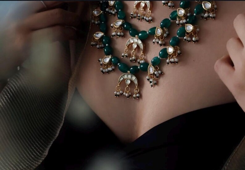 Op de afbeelding: Een gelaagde ketting met groene kralen, gouden accenten en parel-achtige druppels. De ketting heeft een halve maan hanger met een witte steen in het midden.