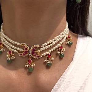 Op de afbeelding: Een meerstrengs parelketting met goudkleurige halve maan hangers, robijnrode accenten en groene edelsteen druppels. De ketting is gelaagd en heeft kleine parel- en edelsteen bungelende bedels.