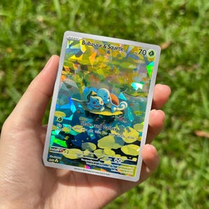 May include: A holographic trading card featuring Bulbasaur and Squirtle. The card has a vibrant, colorful background with the text "Bulbasaur & Squirtle" and "Shake and Gather". The card is held against a blurred green background.