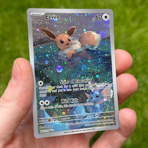 May include: A holographic Eevee trading card with 60 HP, featuring a brown Eevee swimming in water and a blue Vaporeon below. The card text includes Signs of Evolution and Wild Kick attacks, with a 052/072 set number and a silver border design.