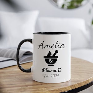 May include: White ceramic coffee mug with a black rim and handle. The mug has a black and white design of a mortar and pestle with the text "Amelia Pharm D Est. 2024".