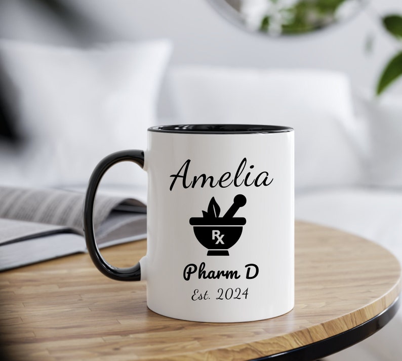 May include: White ceramic coffee mug with a black rim and handle. The mug has a black and white design of a mortar and pestle with the text "Amelia Pharm D Est. 2024".