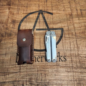 May include: A silver multi-tool with the text "Leatherman Wave" on the side. The multi-tool is closed and sitting on a wooden surface. A brown leather case is next to the multi-tool.