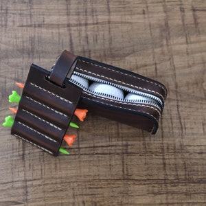 Puede incluir: Un soporte de bolas y tees de golf de cuero marrón con una bolsa con cremallera para bolas de golf. La bolsa está abierta y contiene cuatro bolas de golf blancas. El soporte tiene cuatro ranuras para tees, tres de las cuales están llenas de tees naranja y verde.