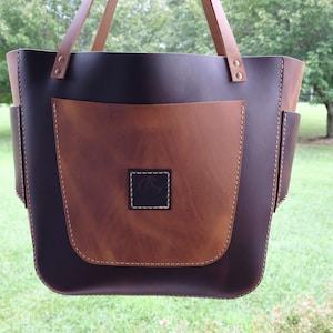 Op de afbeelding: Een bruine leren tote bag met een grote voorzak en twee kleinere zijzakken. De tas heeft een bruine leren band en een zwart leren lichaam. De tas heeft een klein vierkant logo op de voorzak.