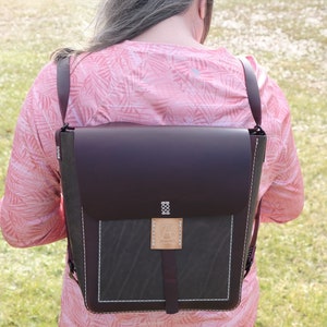 May include: A brown leather backpack with a dark green canvas flap and white stitching. The backpack has a brown leather strap and a small brown leather patch with a logo on the front.