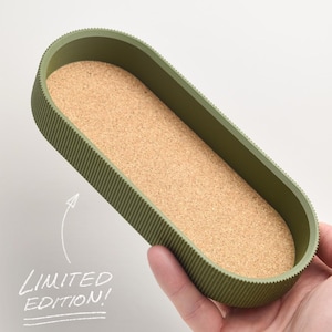 May include: An olive green, oval-shaped tray with a textured exterior and a cork interior. The tray is held in a hand. The text "LIMITED EDITION!" is written in white. This tray is suitable for home decor.
