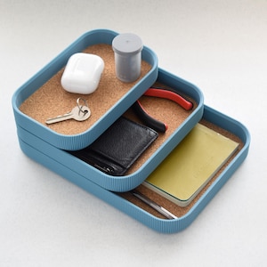 May include: Three stacked blue rectangular trays with cork inserts. The top tray holds a white wireless earbud case, a grey container, and a key. The middle tray contains a black wallet and red pliers. The bottom tray has a yellow notebook and a pen.