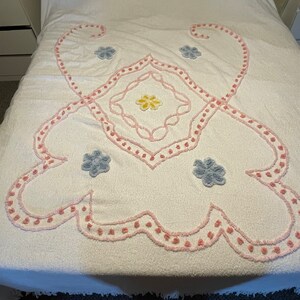 Puede incluir: Un cubrecama blanco con un diseño floral rosa y blanco. El diseño presenta una forma de diamante central con cuatro flores más pequeñas en las esquinas. Las flores son de color azul claro.