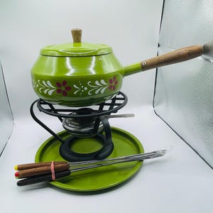 Può includere: Un set per fonduta verde vintage con un design floreale. Il set include una pentola verde con manico in legno, un supporto in metallo nero, un piatto verde e forchette per fonduta. La pentola ha un pomello in legno sul coperchio.