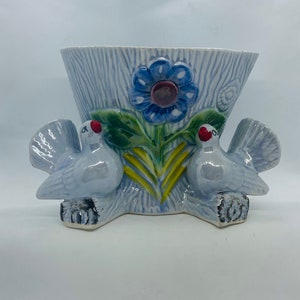 May include: A blue ceramic planter with two white doves on either side. The planter has a flower with a blue centre and a maroon centre. The planter has a textured surface.