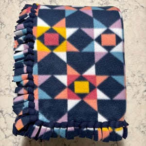 May include: A navy blue fleece blanket with a geometric pattern in pink, yellow, white, and blue. The blanket has a knotted fringe border.